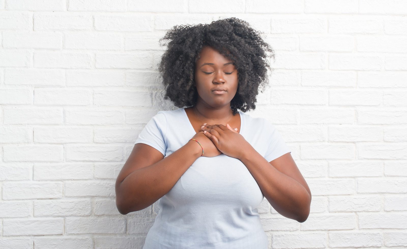 Young african american plus size woman over white brick wall smiling with hands on chest with closed eyes and grateful gesture on face. Health concept.
