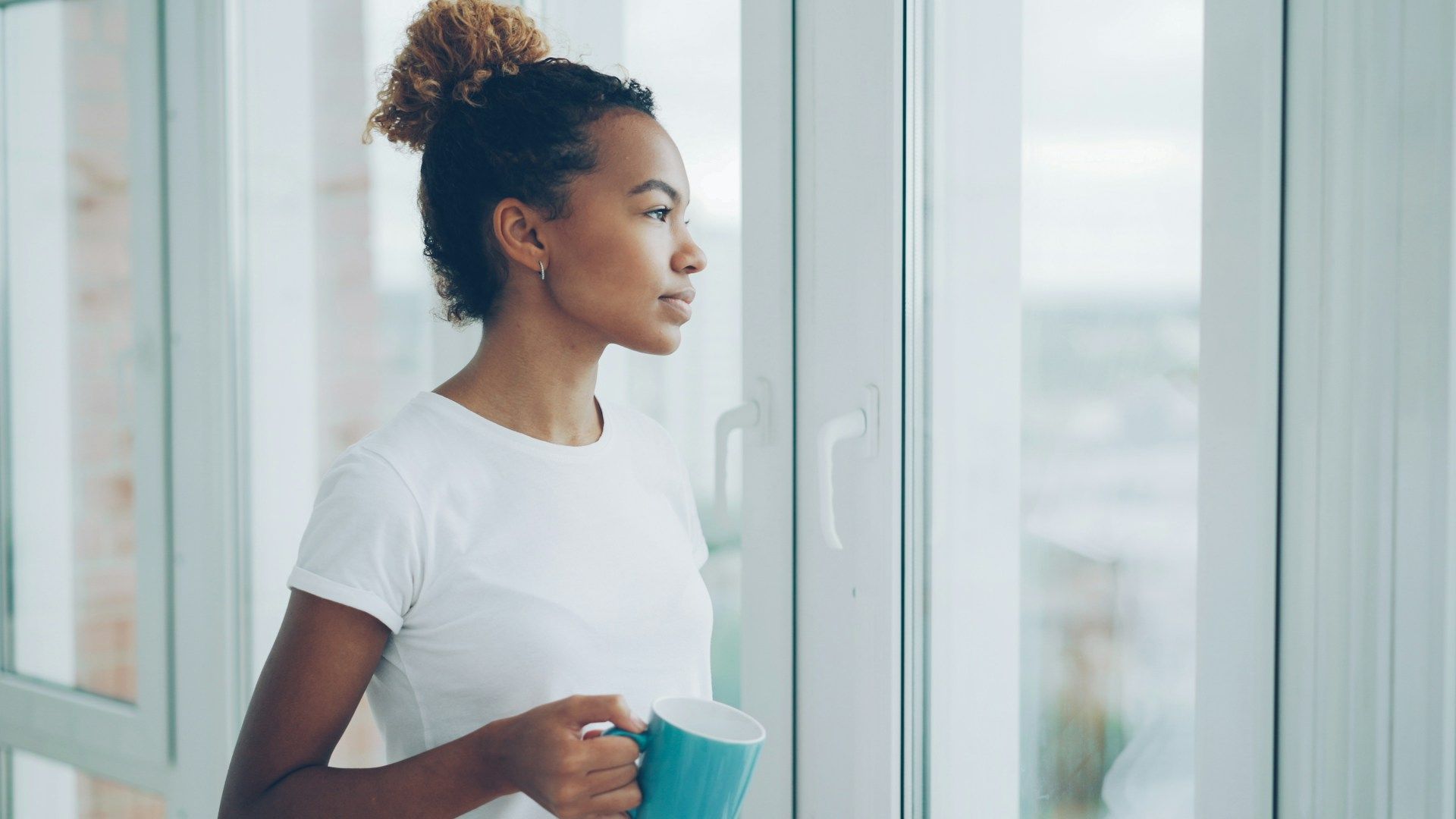 Woman holding a mug and looking out a bright window, quiet morning